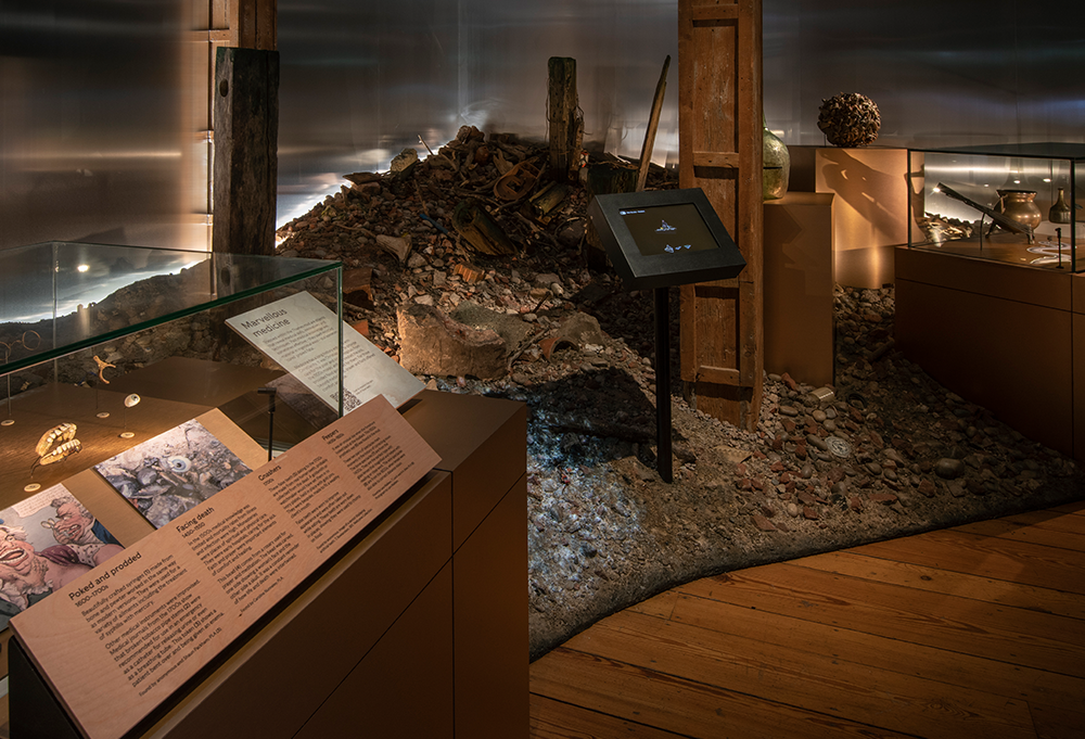 A photo of the exhibition showing the digital interactives mounted on poles. The poles elevate the touchscreens above the sloping, gravel textured setworks which are designed to look like the foreshore of the Thames.