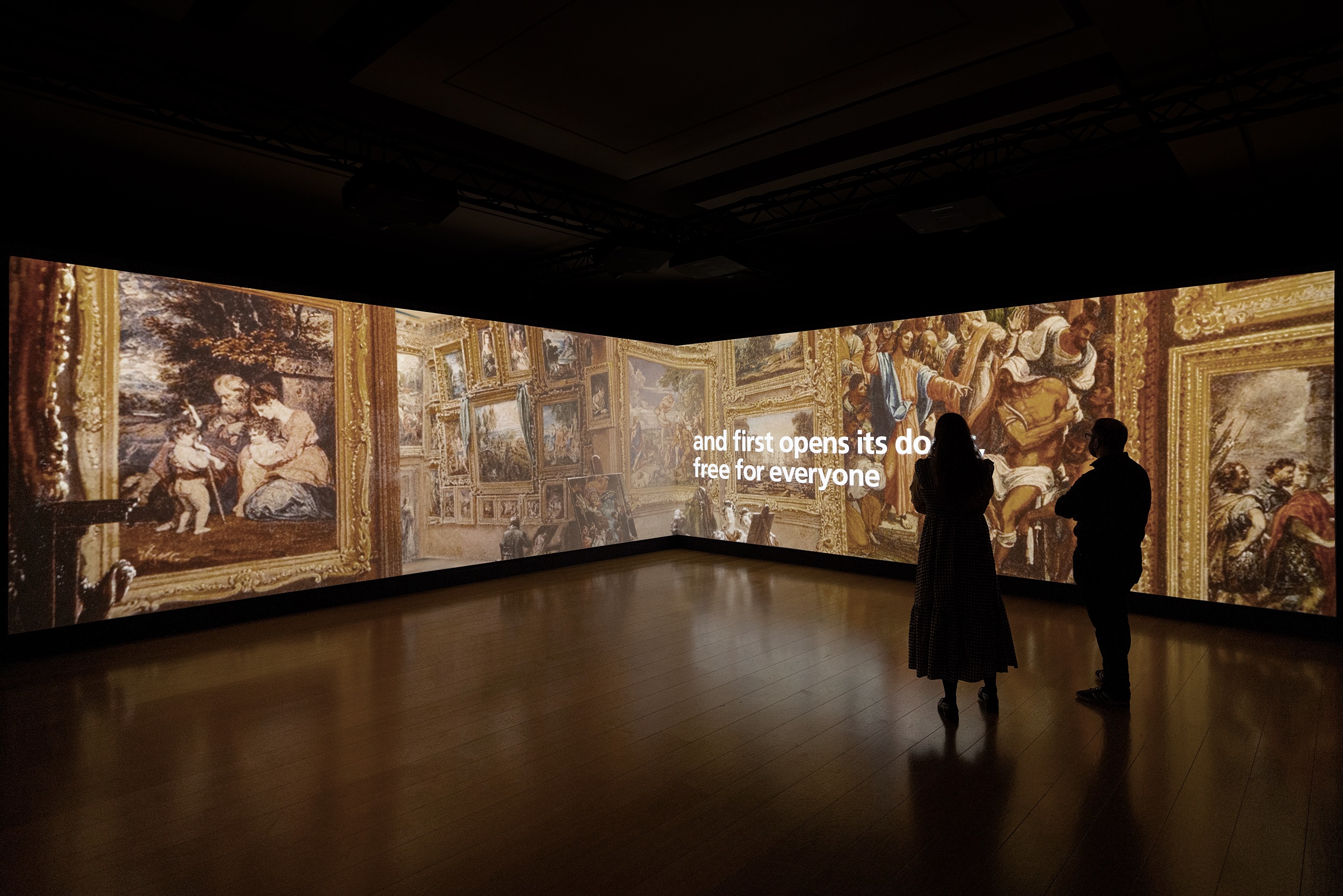 A photo of a large L-shaped video wall displaying the painting The National Gallery when at Mr J. J. Angerstein's House, Pall Mall by Frederick Mackenzie. The painting depicts the interior of the National Gallery in its original location on Pall Pall. Three people stand in front of the screen watching the animation.