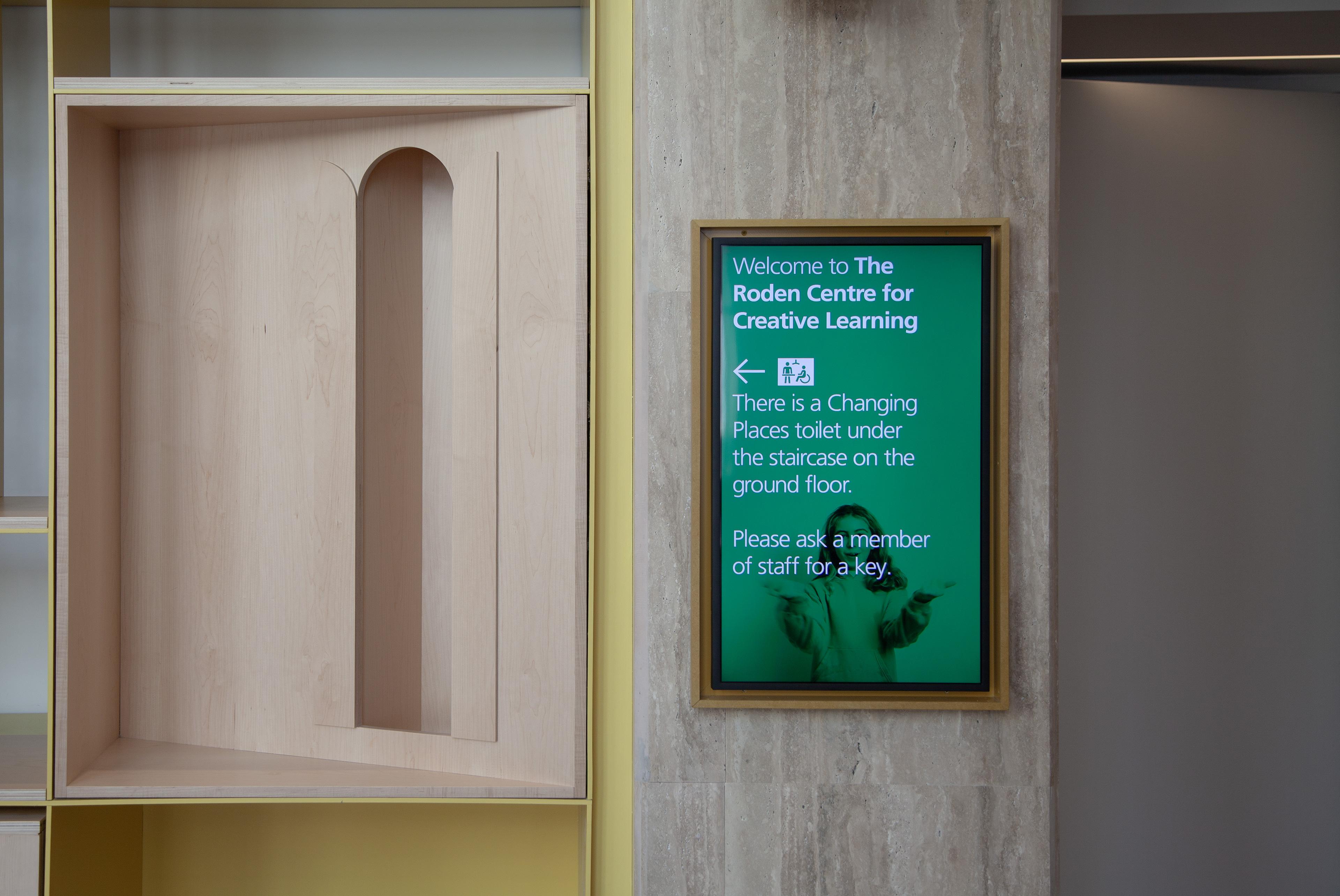 A photo of portrait mounted digital signage screen inside the Roden Centre for Creative Learning at the National Gallery. On the screen is video of a child saying welcome in makaton. The video is overlayed with a welcome message in white text.