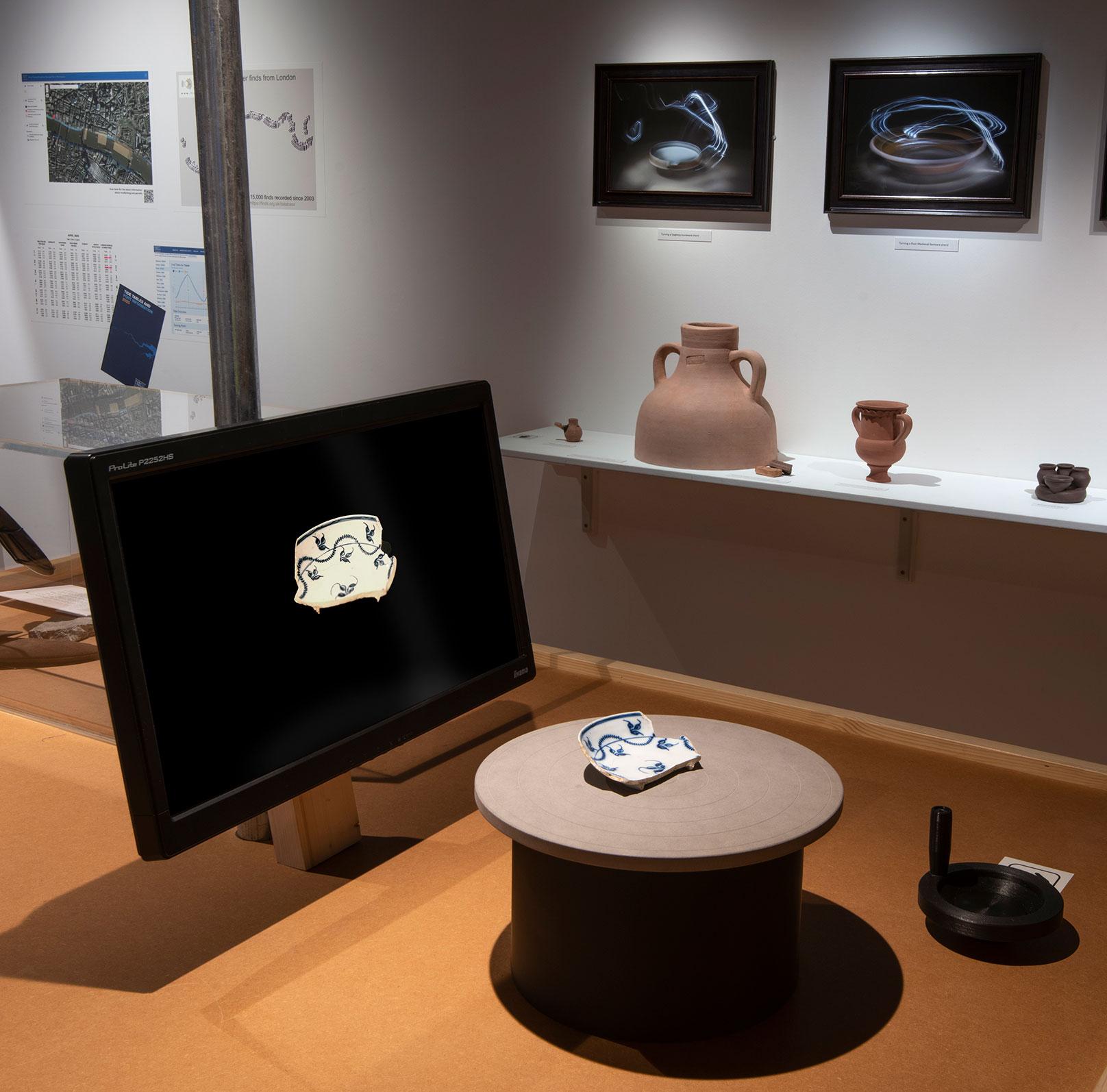 A photograph of an interactive turntable at the Secrets of the Thames exhibition and London Museum. A turntable sits in front of a screen. On top of the turntable is a potter sherd. A handle is in front of the turntable. When visitors turn the handle the image in the screen responds by animating the sherd spinning, using a long exporure effect to show the pottery sherd as a complete rotation and revealing its form 