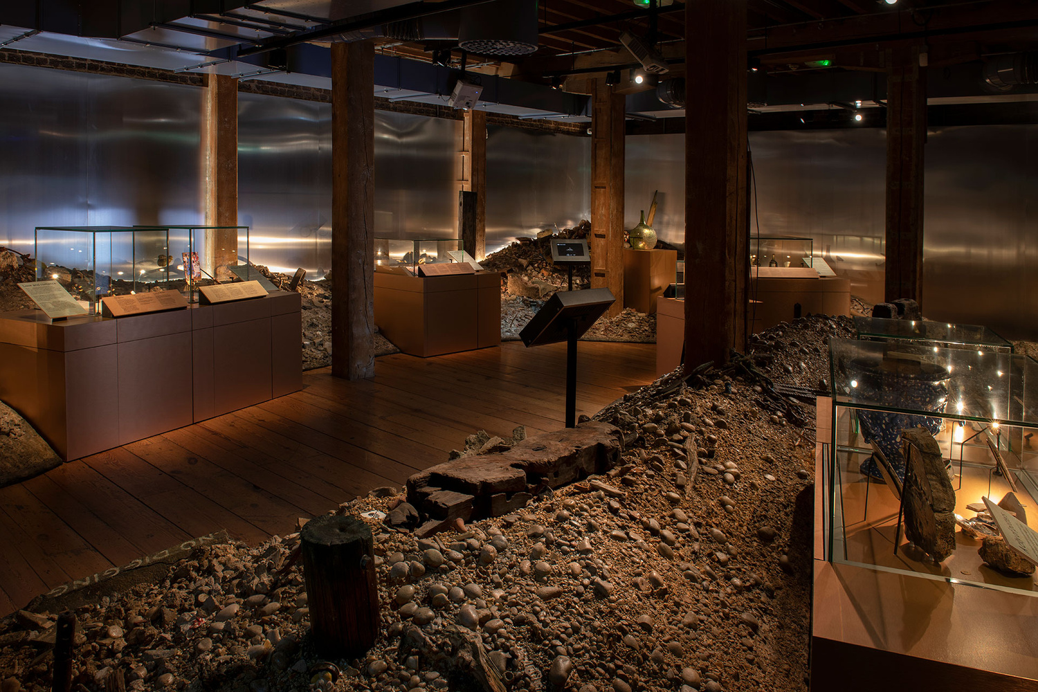A photo of the Secrets of the Thames exhibition. Object show cases and interactive screens on poles protrude from the recreated Thames foreshore in the space.