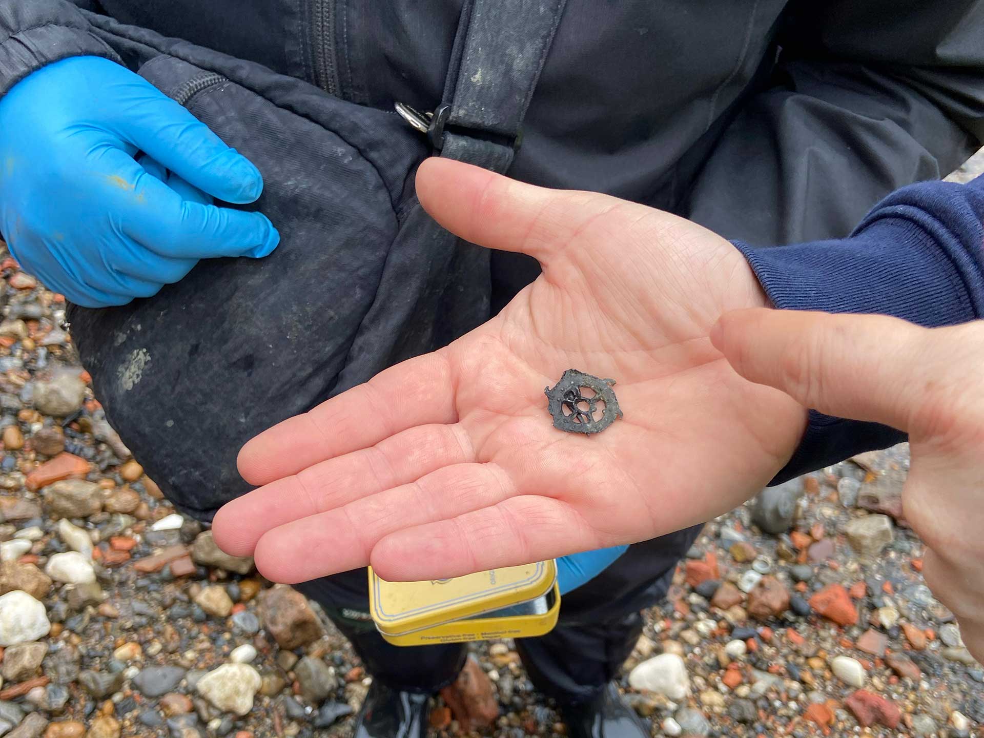 A photo of a hand holding a very small piece of metal shaped like a cart wheel with a hole in the middle, found on the foreshore of the river Thames