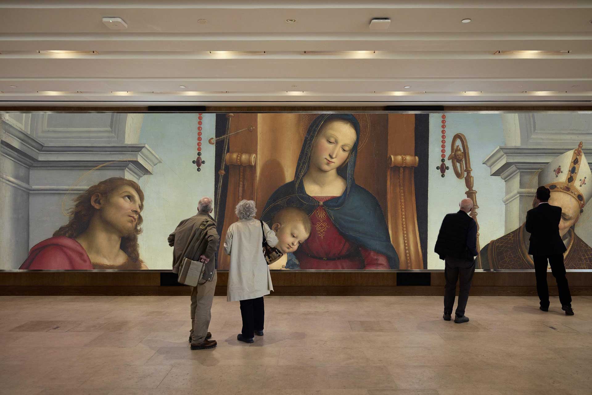 A photograph of the 10 metre wide, 3 metre tall ultra high resolution screen displaying the painting The Ansidei Madonna by Raphael in the National Gallery Sainsbury Wing. Four people are looking at the screen.