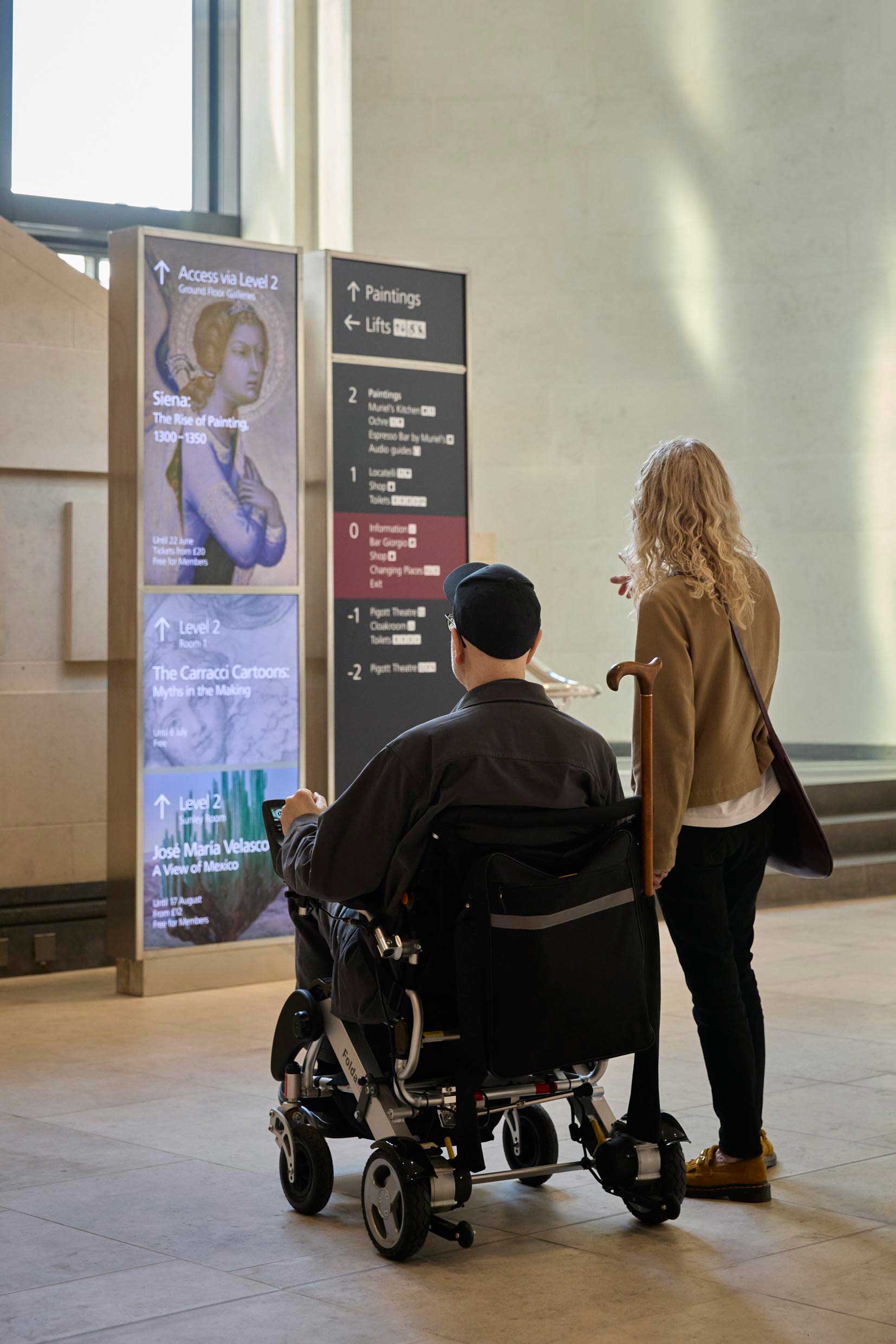 A photograph of a digital wayfinding totem next to the printed wayfinding totem at the bottom of the staircase in the Sainsbury Wing at the National Gallery. A man in a wheelchair and a woman standing next to him are looking at the signage.