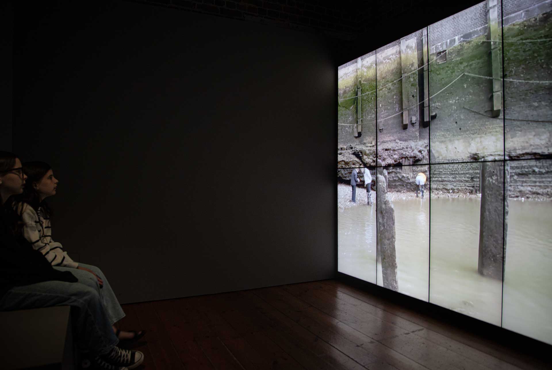 A photo of a video viewing space. 2 girls watch a timelapse film showing mudlarks on the Thames foreshore. The video is played on a large display made up of 8 screen panels.