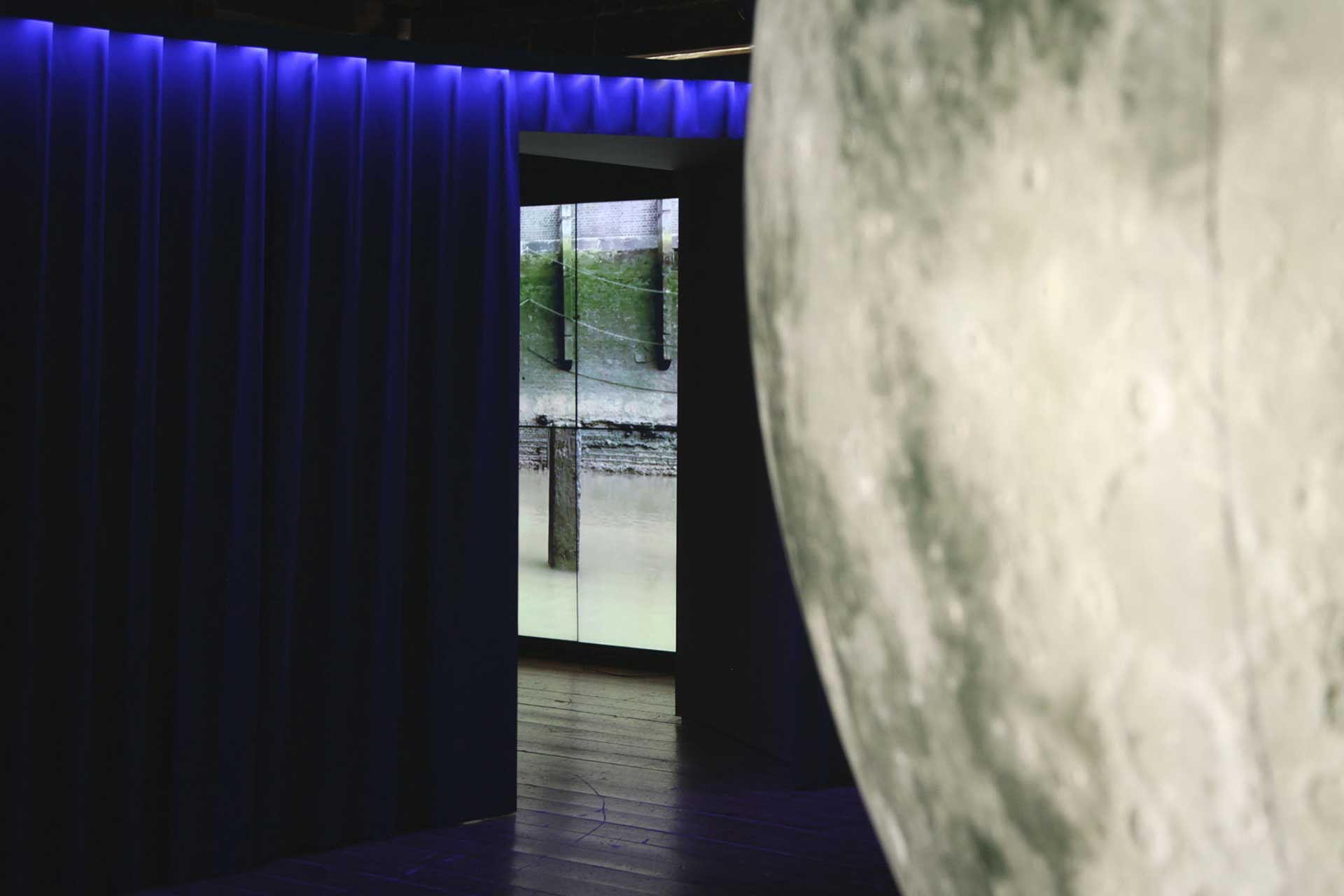 A photo of the entrance tide room viewed from the adjacent room. In the foreground is the large inflatable Moon installation by Luke Jerram. Beyond the moon is a deep blue velvet curtain. Through a gap in the curtain we see a small portion of the tide room video wall 