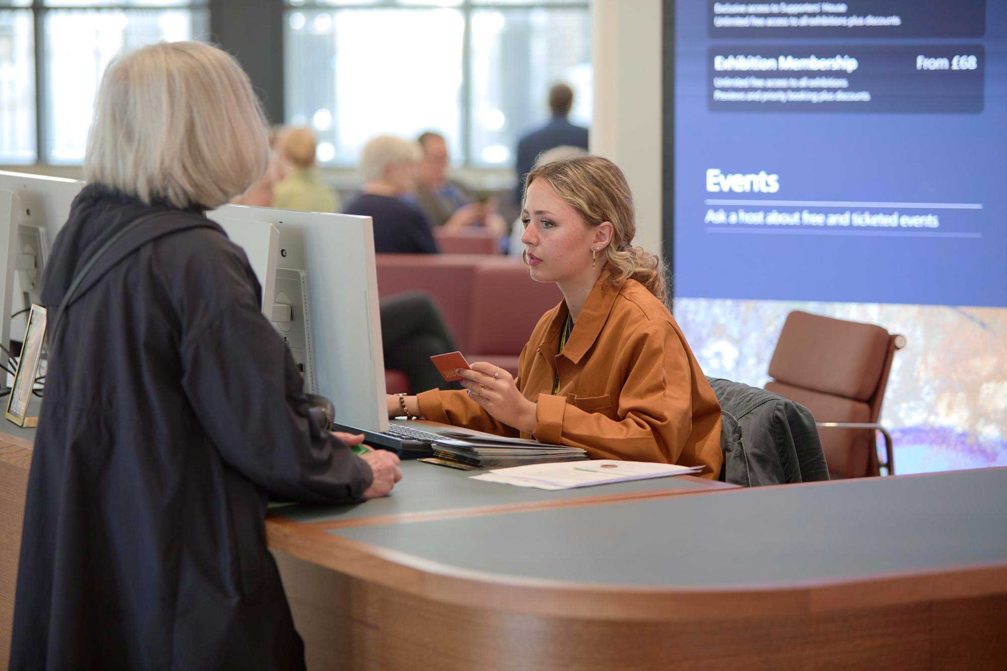 A photograph of the Information desk in the Sainsbury Wing entrance of the National Gallery. A woman sits at the desk helping a visitor with an enquiry. Behind her is a screen 3 metres high and approximately 1.8 metres wide. The screen displays membership and exhibition information and ticket prices.