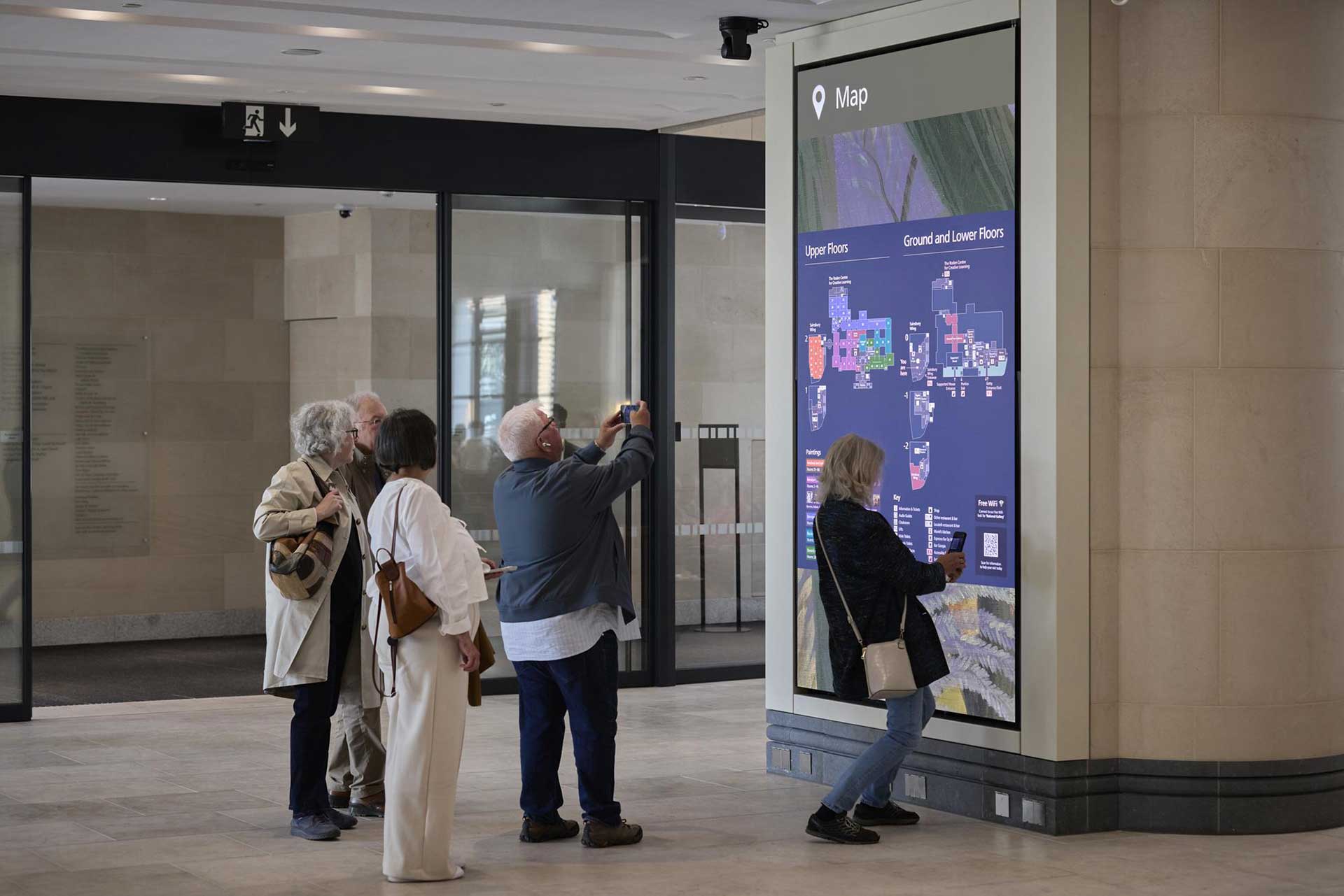 A photograph of the digital map in the Sainsbury Wing entrance in the National Gallery. The screen is 3 metres high and approximately 1.8 metres wide. In front of the screens are several visitors. Two visitors are taking photos of the map with their phones.