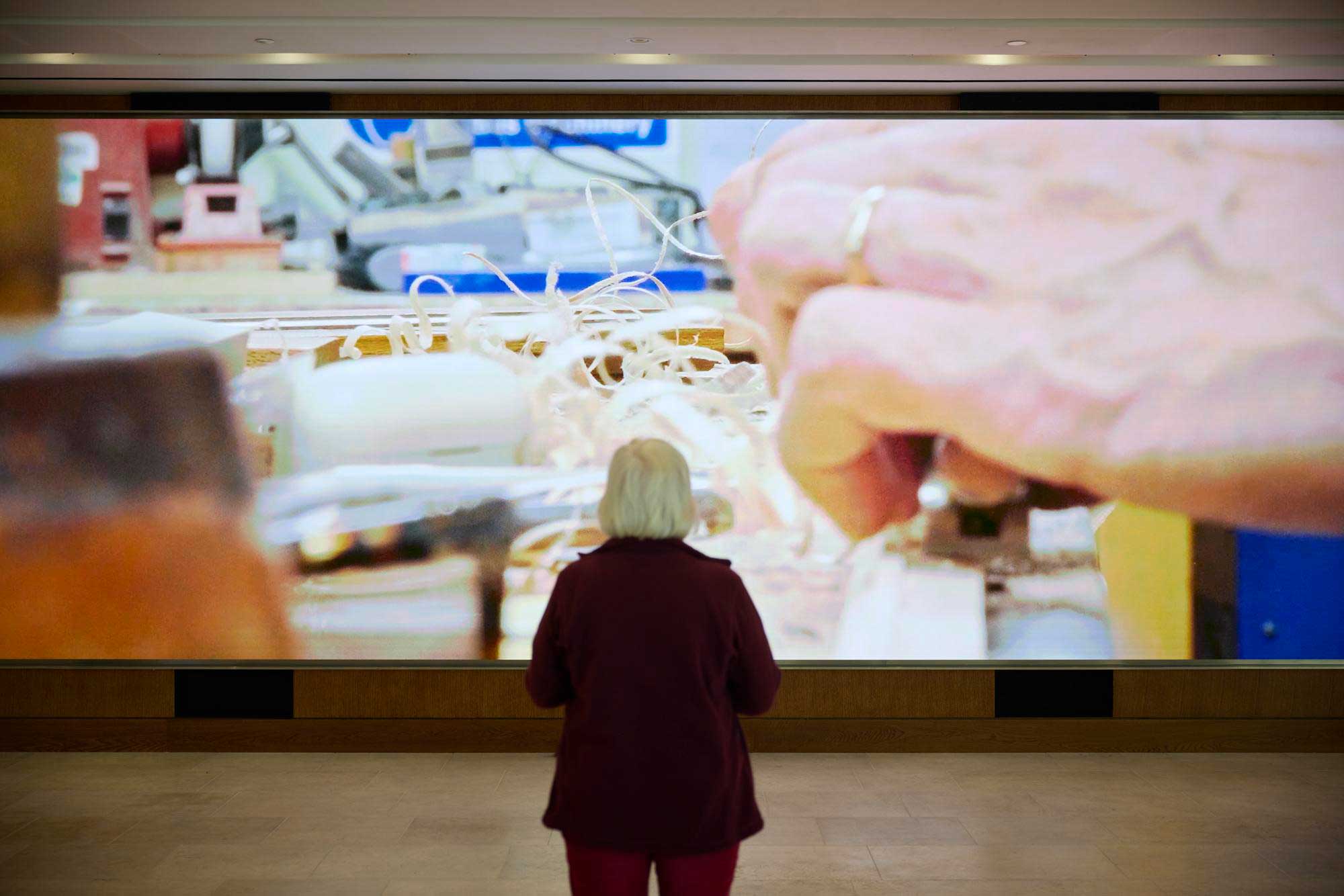 A photograph of the 10 metre wide, 3 metre tall ultra high resolution screen displaying a behind the scenes video in the National Gallery Sainsbury Wing. A woman is standing in front of the screen.