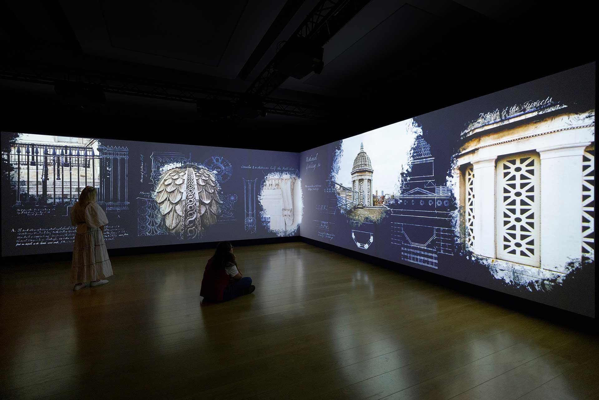 A photo of a large L-shaped video wall displaying the orignal blueprints for the design of the building in Trafalgar Square overlayed with photography of the same building details today. Two people stand in front of the screen watching the animation.