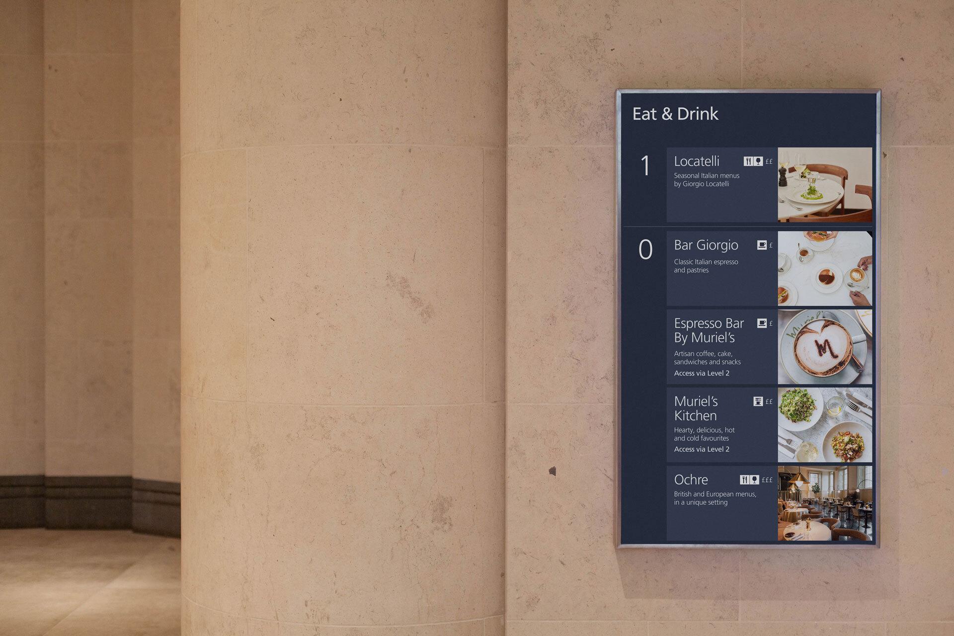 A photograph of a digital screen mounted in portrait orientation on the stone wall inside the Sainsbury Wing at the National Galley. The screen displays a list of places to eat and drink at the Gallery, organised by floor.