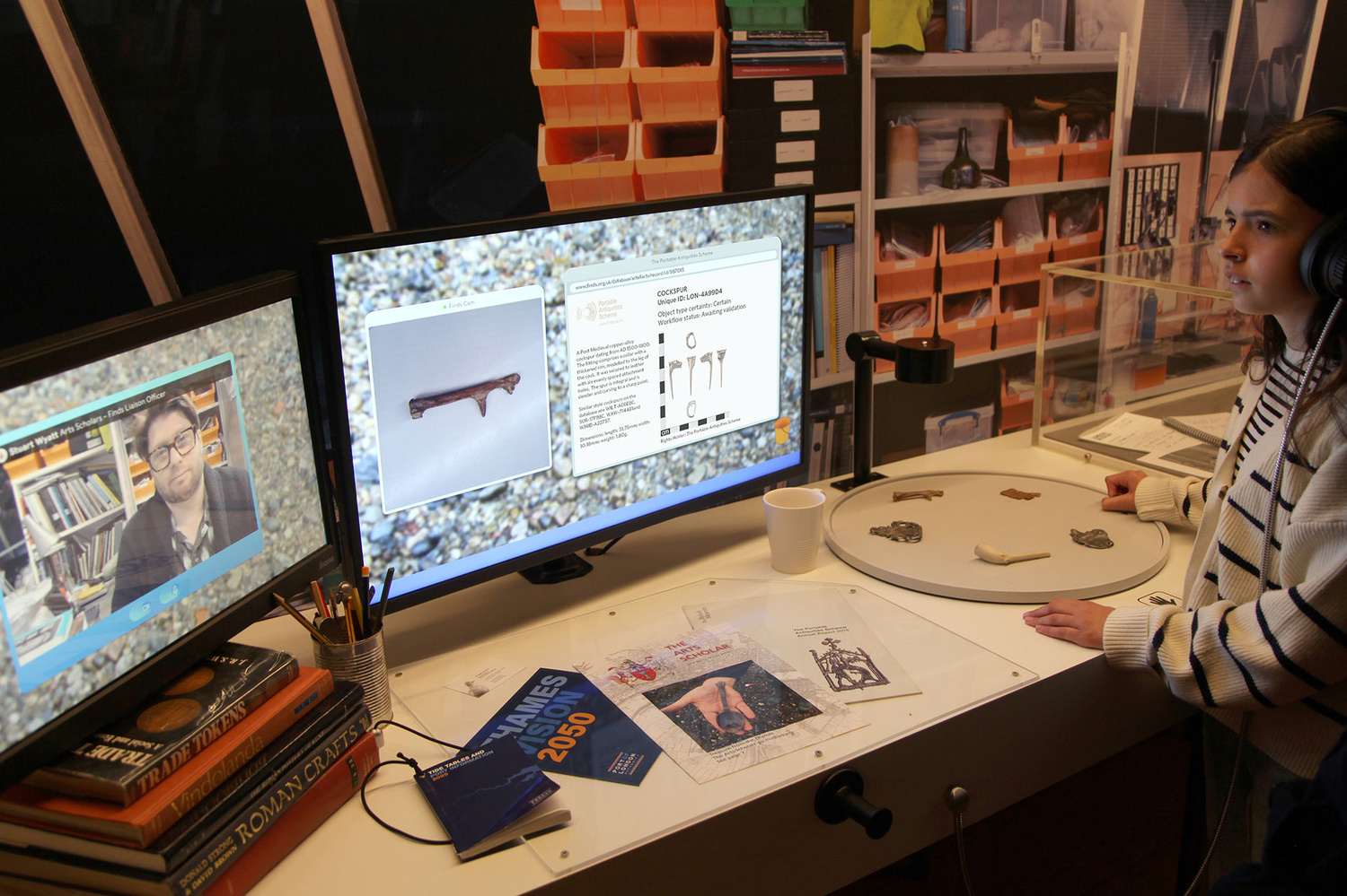 A photograph of an object recognition experience at the Secrets of the Thames exhibition at London Museum Docklands. On a desk are 2 screens side by side, a turntable with 5 objects on it. Above the turntable is a camera. A girl is using the interactive whilst wearing headphones. When the turntable is turned a different object moves under the camera. The computer recognises the object and triggers content on the 2 screens. The left screen plays a video of Finds officer Stuart Wyatt describing the object. The right screen displays the object record in the Portable Antiquities Finds Database. 