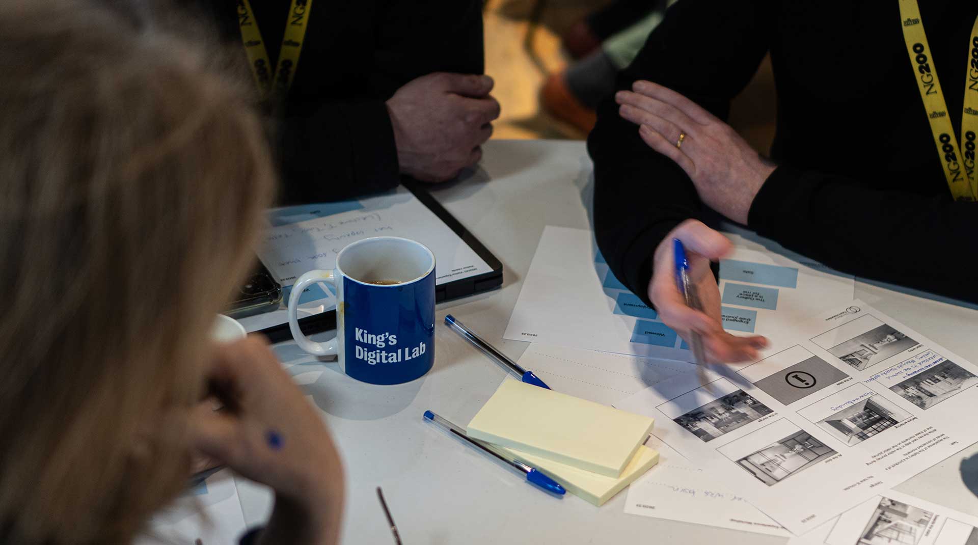 A photo taken during an in-person workshop session at the National Gallery. 3 people sit around a table. On the table are worksheets, post-it notes and pens.