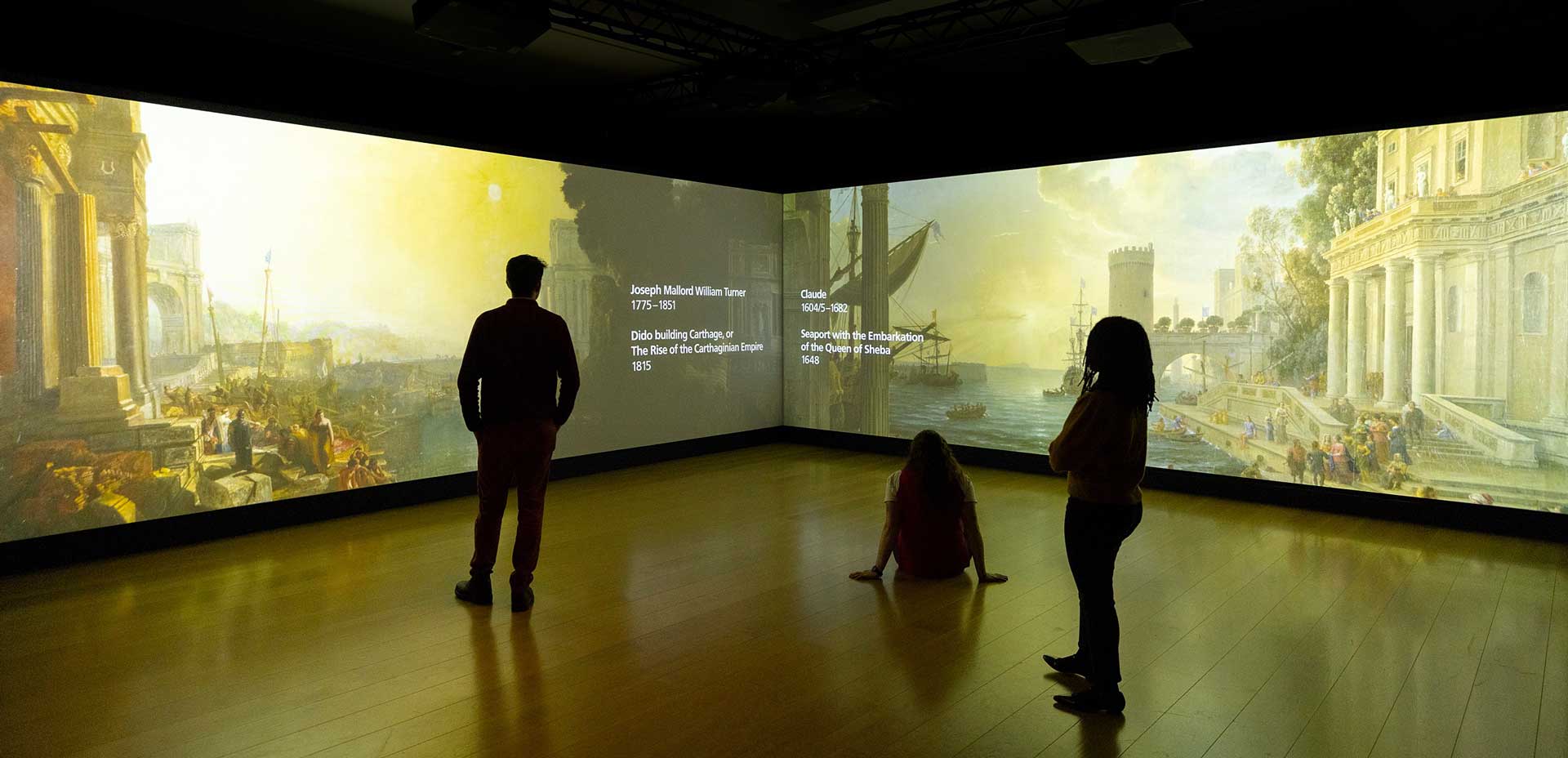 A photo of a large L-shaped video wall displaying the painting Dido building Carthage by J W M Turner on the left and Seaport with the Embarkation of the Queen of Sheba by Claude on the right. Three people stand in front of the screen watching the animation.